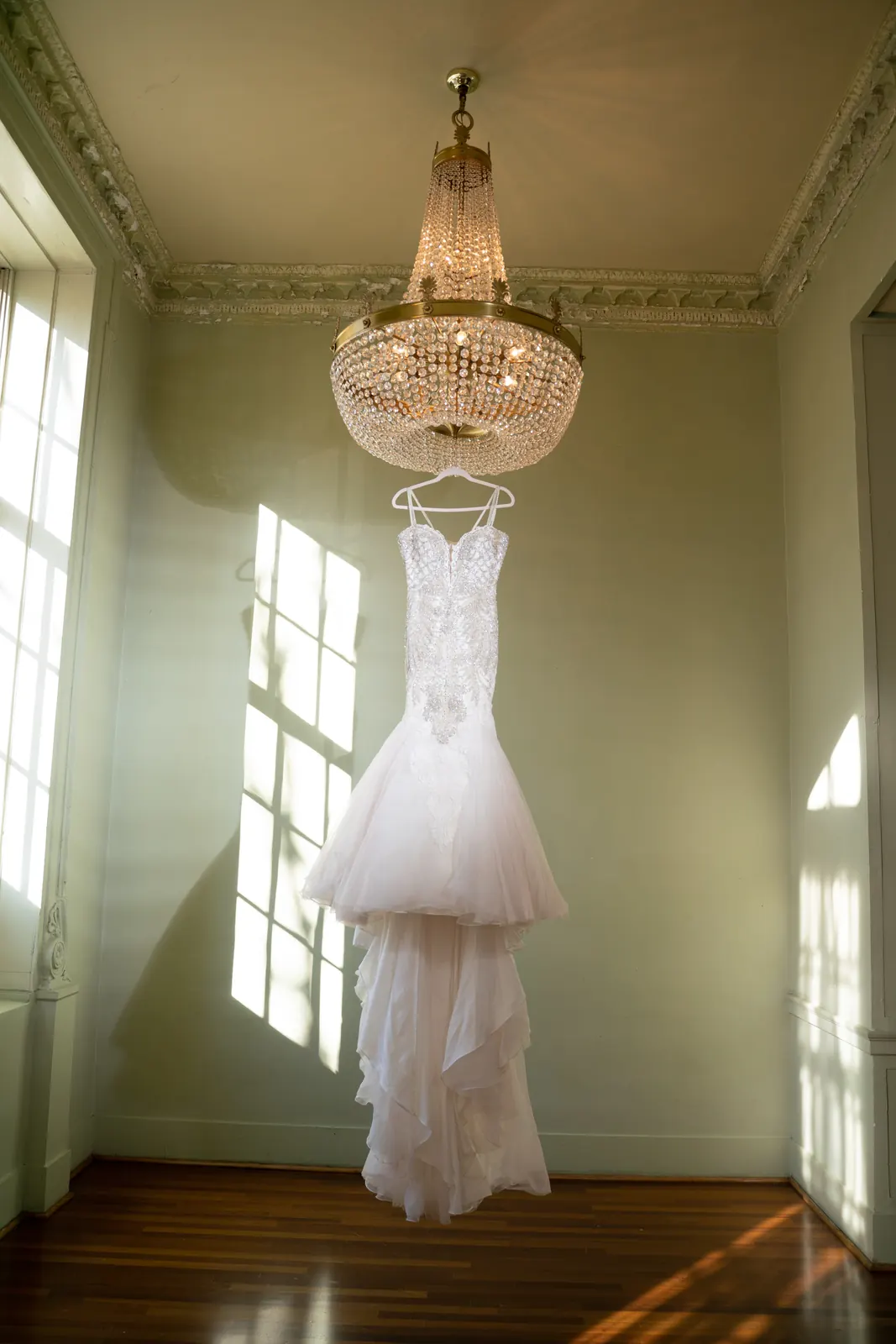 Lace wedding gown hanging from a chandelier in a bright, high-ceilinged room